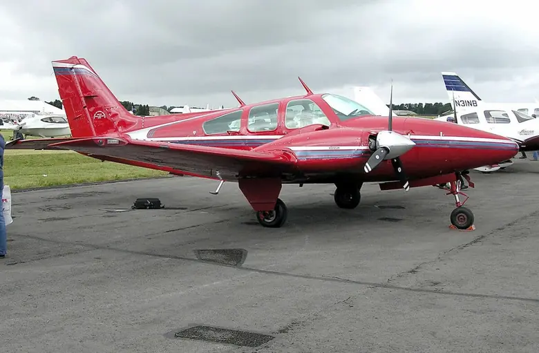 Red and white Beechcraft Baron twin parked on an airport apron with other aircraft behind it