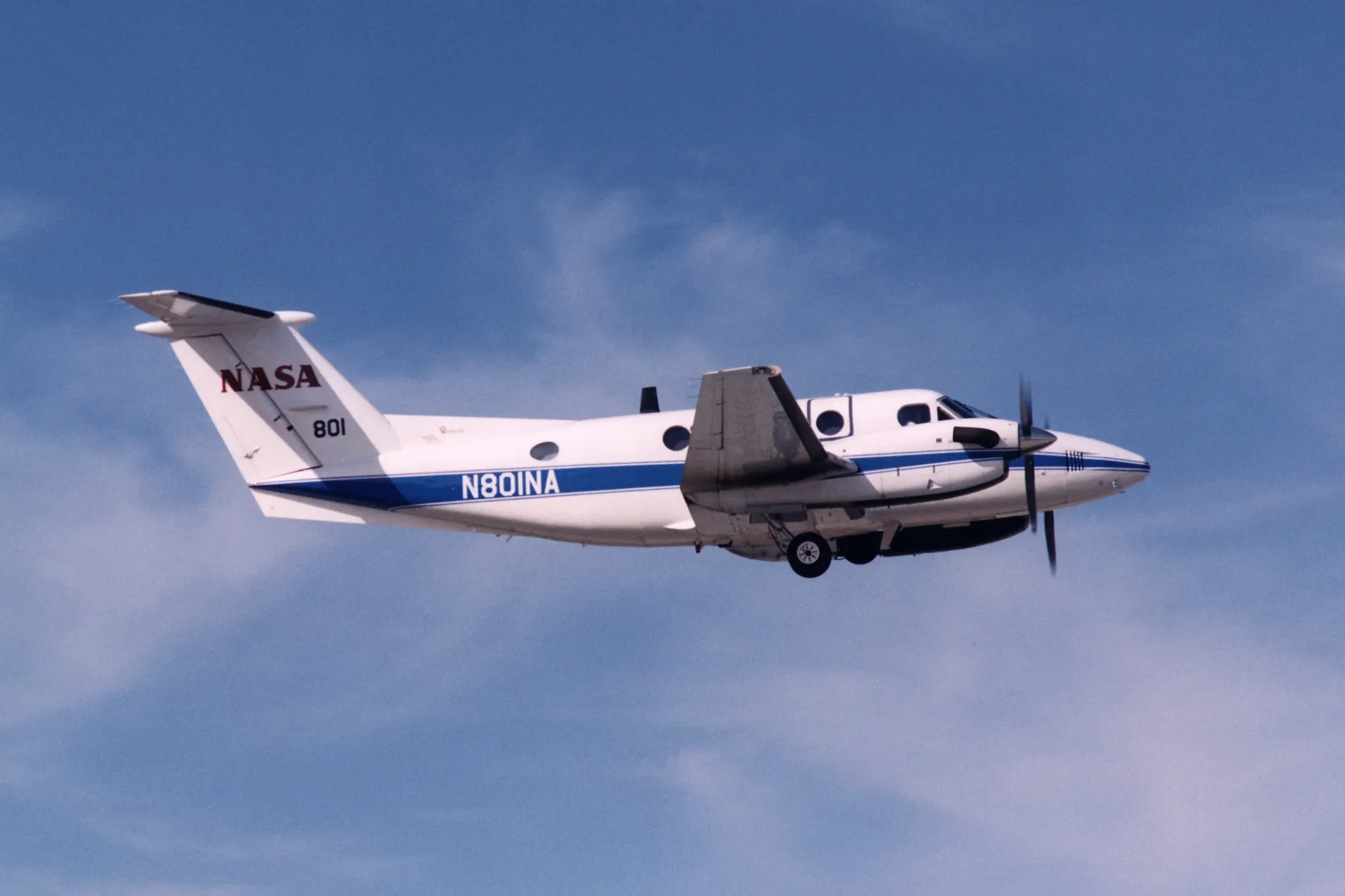 A white and blue twin-turboprop King Air banking away just after takeoff against a blue sky with thin cloud