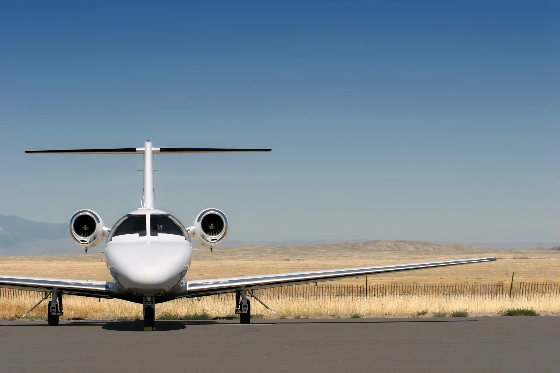 A white twin-engine business jet seen head-on on an airport apron with dry hills behind