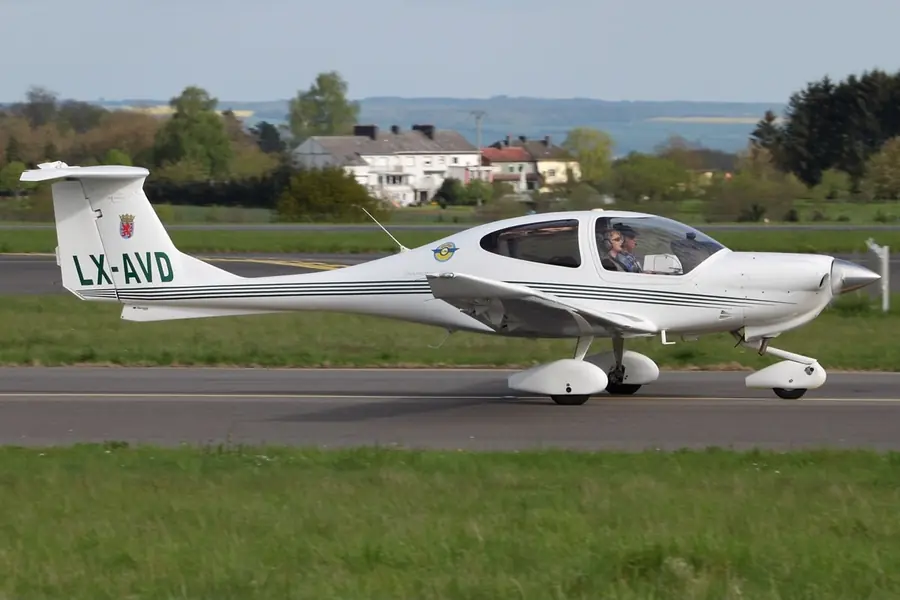 White Diamond DA40 with a T-tail taxiing on a paved strip beside a grass field with trees and houses behind