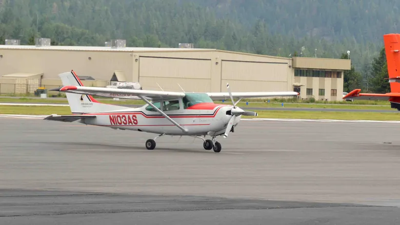 Cessna 182 with a red stripe parked beside an orange Quest Kodiak on an airport apron with forested hills behind