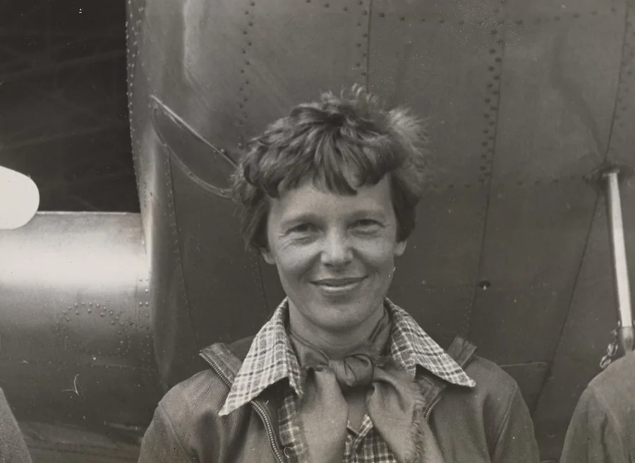 Amelia Earhart smiling in a leather jacket and plaid scarf, standing under the nose of her Lockheed Electra in 1937