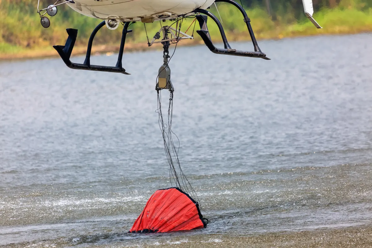 A helicopter lowering a red collapsible bucket into a lake to refill with water